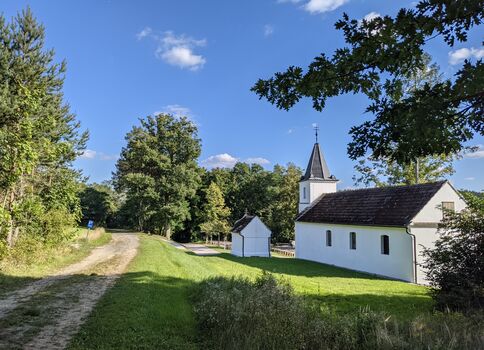 Ländliche Szene: Ein weißes Kirchengebäude mit Turm steht neben einem kleinen weißen Nebengebäude auf grüner Wiese. Ein unbefestigter Weg schlängelt sich durch die links verlaufende Baumallee, während der klare blaue Himmel mit wenigen Wolken zu sehen ist.