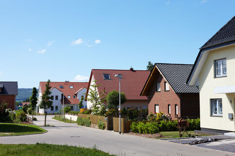 Blick auf eine ruhige Wohnstraße mit modernen Einfamilienhäusern, roten Ziegeldächern und gepflegten Vorgärten. Die Straße windet sich zwischen Häusern entlang, im Hintergrund blauer Himmel. Eine Straßenlaterne steht am Rand, einige Bäume und Sträucher säumen den Gehweg.