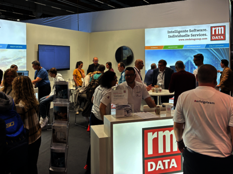 Auf einer Messehalle: Ein RM-DATA-Stand mit leuchtendem Logo, von vielen Besuchern umgeben. Informationsmaterial liegt auf Tischen aus; Banner zeigen „Intelligente Software. Individuelle Services“ im Hintergrund.