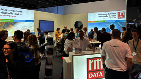 Auf einer Messehalle: Ein RM-DATA-Stand mit leuchtendem Logo, von vielen Besuchern umgeben. Informationsmaterial liegt auf Tischen aus; Banner zeigen „Intelligente Software. Individuelle Services“ im Hintergrund.
