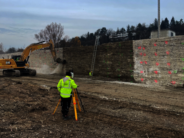 A construction site with a yellow CAT excavator digging near a stone retaining wall, while a worker in a neon jacket sets up a surveying tripod. A ladder leans on the wall; the ground is dirt.