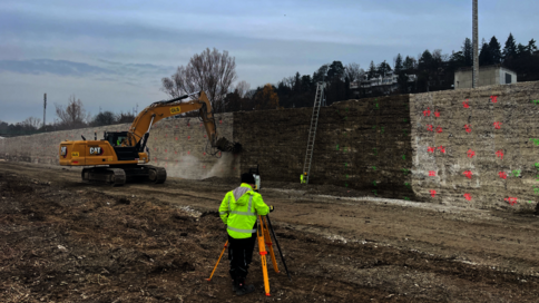 Auf einer Baustelle gräbt ein gelber Raupenbagger Erde neben einer Steinmauer; ein Bauarbeiter in Leuchtwarnjacke bedient ein Vermessungsgerät auf einem Stativ, während eine Leiter an der Wand lehnt.