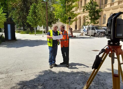 Zwei Vermesser in Warnwesten prüfen gemeinsam einen Bauplan auf einem sonnigen Platz vor einem historischen Gebäudekomplex. Im Vordergrund steht ein Vermessungsgerät auf einem Stativ; dahinter wachsen Bäume, rechts ist ein großes Gebäude zu sehen.