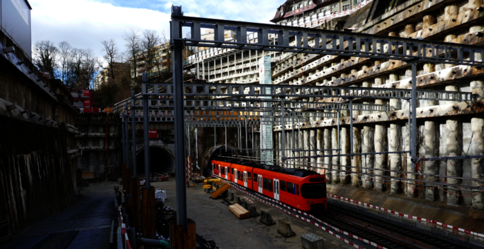 Eine Baustelle an einer Eisenbahnstrecke: Ein roter Passagierwagen steht auf Gleisen in einem Bauareal mit Stahlrahmen, Holzstützen und Steinwänden. Im Hintergrund ist ein Tunnelzugang sichtbar; der Himmel ist blau mit Wolken.
