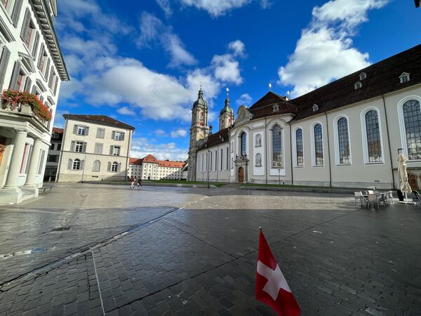 Breite Aufnahme eines europäischen Stadtplatzes: links farbige Häuser mit Blumenkästen, in der Mitte eine weiße Kirche mit zwei Türmen, rechts weitere historische Fassaden; im Vordergrund liegt ein kleines rotes Schweizer Fähnchen auf dem gepflasterten Platz; blauer Himmel mit Wolken.