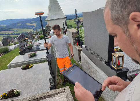 Zwei Arbeiter arbeiten auf einem Friedhof: Einer hält ein langes Messwerkzeug, der andere schaut auf ein Smartphone- oder Tablet-Display, vermutlich zur Dokumentation. Im Hintergrund sind Grabsteine und eine grüne Landschaft sichtbar.