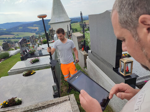 Zwei Arbeiter arbeiten auf einem Friedhof: Einer hält ein langes Messwerkzeug, der andere schaut auf ein Smartphone- oder Tablet-Display, vermutlich zur Dokumentation. Im Hintergrund sind Grabsteine und eine grüne Landschaft sichtbar.