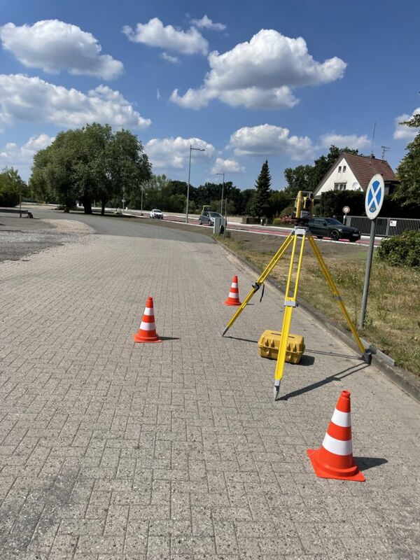 Roadwork scene on a suburban street with orange traffic cones and a yellow surveying tripod; cars are parked along the road, with a house and trees in the background under a bright blue sky.