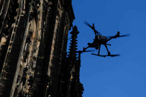 Eine Drohne schwebt vor der Fassade einer gotischen Kathedrale mit kunstvollen Steinmetzarbeiten gegen einen klaren blauen Himmel.