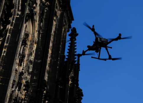 Eine Drohne schwebt vor der Fassade einer gotischen Kathedrale mit kunstvollen Steinmetzarbeiten gegen einen klaren blauen Himmel.