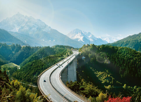 Eine kurvige Autobahnbrücke windet sich über eine grüne Berglandschaft. Bewaldete Hügel umgeben das Tal, dahinter ragen schneebedeckte Berggipfel empor. Die Szene ist sonnig und klar.