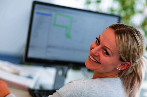 Eine lächelnde Frau sitzt an einem Schreibtisch vor einem Computerbildschirm, der eine grafische Softwareoberfläche mit grünem Layout zeigt. Die Szene wirkt wie ein Büroarbeitsplatz; die Frau schaut in die Kamera.