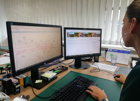 Eine Person sitzt an einem Schreibtisch mit zwei Monitoren: links eine technische Skizze, rechts eine Browser-Seite. Tastatur, Maus und Büro-Utensilien liegen vor ihr; Fensterjalousien im Hintergrund.