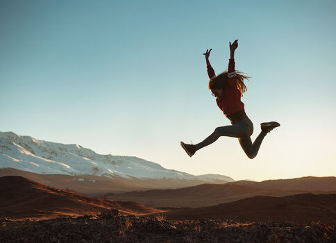 Une personne en tenue de sport rouge saute en hauteur dans un paysage désertique et montagneux, au soleil bas, avec des montagnes enneigées à l'arrière-plan.