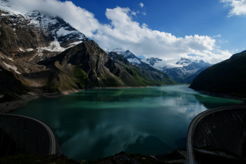 Türkisfarbener Bergsee in einem engen Alpental, umgeben von schroffen, schneebedeckten Gipfeln. Die Wasseroberfläche spiegelt Himmel und Wolken wider; im Vordergrund sind Bogenstrukturen eines Damms sichtbar.