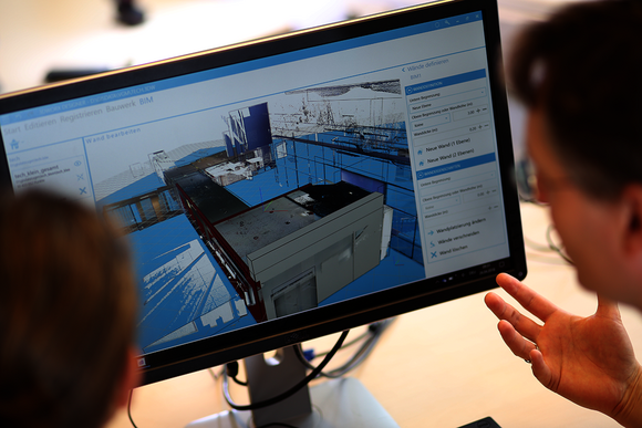 Zwei Personen betrachten einen Monitor mit einer 3D-CAD-Darstellung einer Industrieanlage in Blau, inklusive Maschinen und Strukturen; es wirkt wie eine technische Bauplanung.