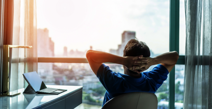 Eine Person sitzt entspannt auf einem Stuhl vor einem großen Fenster und blickt auf die Stadt. Die Arme sind hinter dem Kopf verschränkt. Auf dem Schreibtisch daneben liegt ein Laptop, während das Tageslicht durch die Vorhänge fällt.