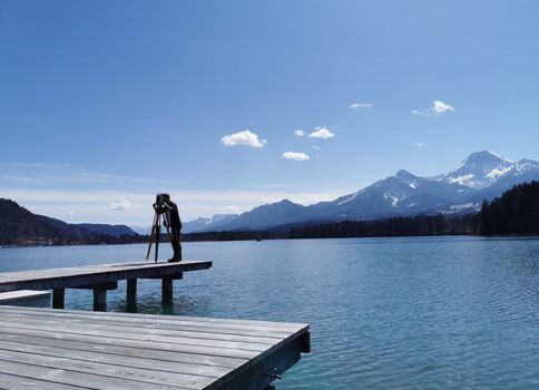 Ein Fotograf steht mit Stativ am Rand eines Holzstegs, der in einen stillen See führt; im Hintergrund erstreckt sich eine Berglandschaft mit schneebedeckten Gipfeln unter blauem Himmel.
