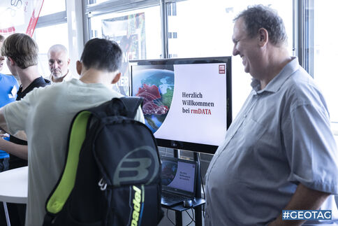 Mehrere Besucher stehen an einem Messestand. Im Mittelpunkt ist ein Monitor zu sehen mit der Aufschrift "Herzlich Willkommen bei rmDATA". Eine Person mit neon-grünem Rucksack betrachtet den Bildschirm, rechts lächelt ein Mann in hellblauem Hemd; weitere Besucher im Hintergrund.