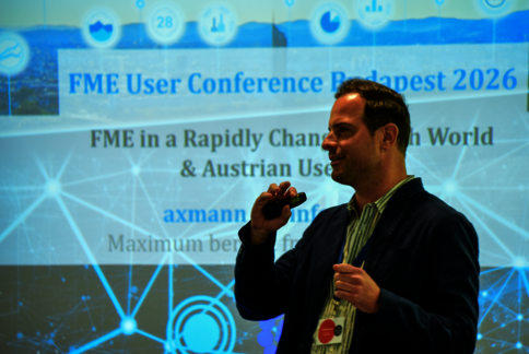 Ein Mann hält ein Mikrofon vor einem Vortrag bei der FME User Conference Budapest 2026. Im Hintergrund eine Projektionswand mit dem Konferenztitel und digitalen Netzwerkgrafiken.