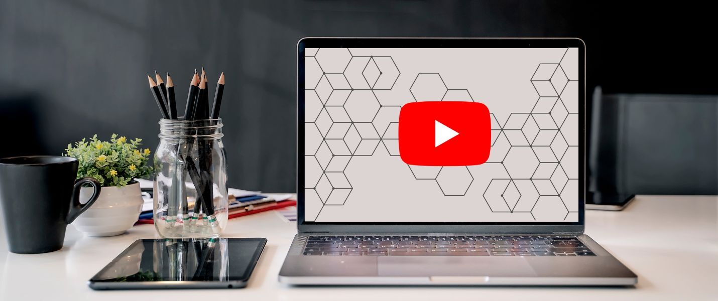 Moderner Arbeitsplatz: Ein Laptop mit YouTube-Logo auf dem Bildschirm steht auf einem Schreibtisch; daneben liegen ein Tablet, eine Tasse Kaffee, ein Glas mit Stiften, eine Pflanze und ein Notizbuch.