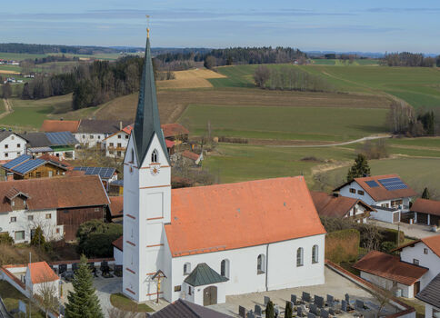 Vue aérienne d'un petit village rural centrée sur une église blanche au toit rouge et à son clocher élancé, entourée de maisons à tuiles rouges et de vastes champs verdoyants.