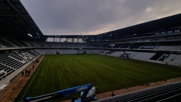 An empty stadium with a green grass field in the center, surrounded by seating stands and a running track around the pitch; a few people walk along the track under a cloudy sky.