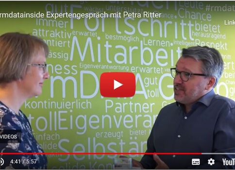 Deux interlocuteurs, une femme à gauche et un homme à droite, discutent face à face dans un cadre professionnel; un mur vert en arrière-plan est couvert de mots en allemand liés au travail.