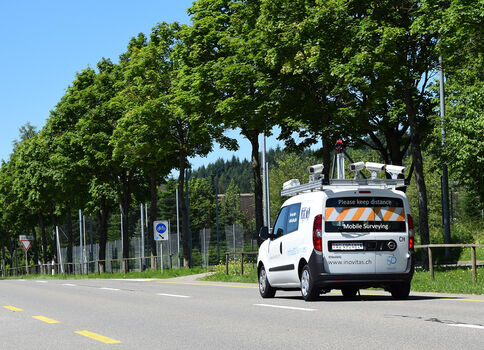 Weißes Messfahrzeug mit Ausrüstung auf dem Dach fährt auf einer zweispurigen Straße entlang einer Baumallee; auf der Heckklappe steht „Mobile Surveying“ und eine Website, am Wagen sichtbar ist der Hinweis „Please keep distance“.