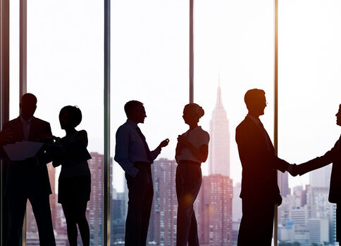 Silhouetten von Geschäftspersonen in Anzügen stehen in einem Büro mit großen Fenstern. Einige tauschen Gespräche, zwei Personen schütteln sich die Hände. Im Hintergrund erstreckt sich eine städtische Skyline bei warmem Licht.