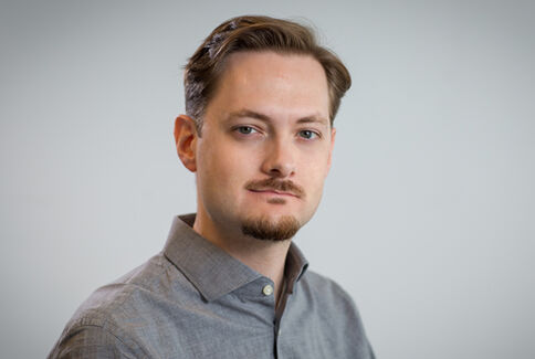 Porträt eines jungen Mannes mit kurzen braunen Haaren und Kinnbart, trägt hellgraues Hemd und schaut in die Kamera; heller, unscharfer Hintergrund.