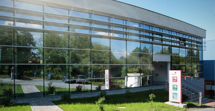 Moderne Glasfassade eines flachen Gebäudes mit umliegendem Grün. Große Fensterscheiben spiegeln Bäume und blauen Himmel. Rechts am Gebäude befindet sich der Eingang mit Treppe und Geländer.