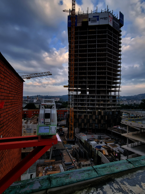 Eine Baustelle in der Stadt mit einem teilweise fertigen Hochhaus aus Stahlbeton; daneben ein Baukran. Im Vordergrund Baugeräte und eine rote Backsteinwand, darüber ein wolkiger Himmel.