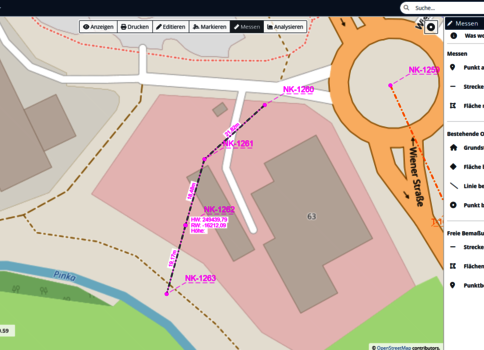 Eine Kartendarstellung eines städtischen Gebietes mit grauen Gebäuden, einem Fluss unten links und einer orangen Umgehungsstraße rechts. Eine magentafarbene Trasse zieht sich durch das Gelände und ist durch mehrere NK-Bezeichnungen markiert, offenbar eine geplante Route.