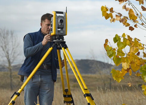 Ein Vermesser richtet ein Totalstation-Vermessungsgerät auf einem gelben Dreibein-Stativ in einer offenen Landschaft mit Herbstbäumen.