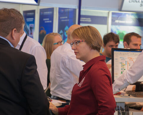 Eine Frau mit Brille in rotem Polo spricht auf einer Messe mit einem Mann im Anzug; im Hintergrund stehen Informationsstände und Monitore.