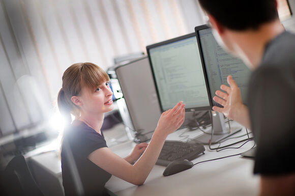 Zwei Personen sitzen an einem Schreibtisch mit mehreren Monitoren. Eine Frau schaut den Gegenüber an und erklärt etwas, während beide auf die Computerbildschirme schauen. Die Szene wirkt wie eine Büro- oder Lernumgebung mit Computern.