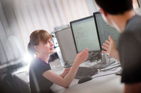 Zwei Personen sitzen an einem Schreibtisch mit mehreren Monitoren. Eine Frau schaut den Gegenüber an und erklärt etwas, während beide auf die Computerbildschirme schauen. Die Szene wirkt wie eine Büro- oder Lernumgebung mit Computern.