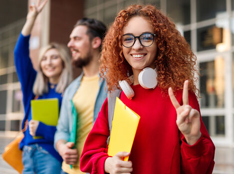 Junge Frau mit rotem Pullover, Brille und Kopfhörern um den Hals lächelt und zeigt das Peacezeichen; sie hält einen gelben Ordner. Im Hintergrund stehen weitere Studenten mit bunten Ordnern vor einem Gebäude.