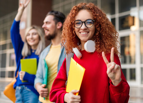 Junge Frau mit rotem Pullover, Brille und Kopfhörern um den Hals lächelt und zeigt das Peacezeichen; sie hält einen gelben Ordner. Im Hintergrund stehen weitere Studenten mit bunten Ordnern vor einem Gebäude.