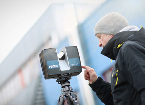 Ein Mann in warmer Kleidung und Mütze bedient ein auf einem Stativ montiertes Laserscanner-Gerät im Freien vor einem Industriegebäude.