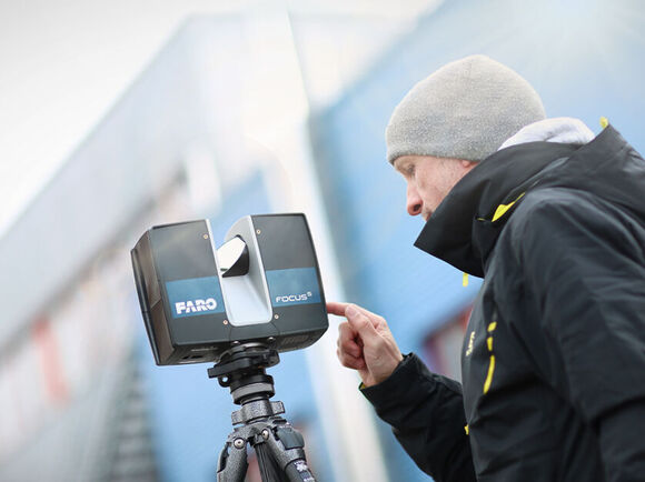 Ein Mann in warmer Kleidung und Mütze bedient ein auf einem Stativ montiertes Laserscanner-Gerät im Freien vor einem Industriegebäude.