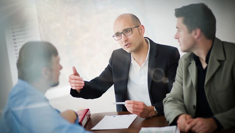 Drei Geschäftsleute sitzen an einem Tisch. Der mittlere Mann mit Brille erklärt etwas und hält ein Dokument, während die beiden anderen zuhören.