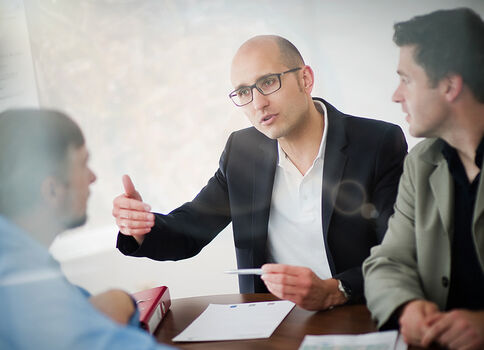 Drei Geschäftsleute sitzen an einem Tisch. Der mittlere Mann mit Brille erklärt etwas und hält ein Dokument, während die beiden anderen zuhören.