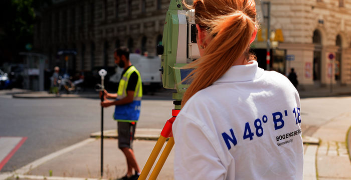Sur une rue urbaine, une technicienne vue de dos ajuste un théodolite sur un trépied; elle porte un polo blanc. En arrière-plan, un collègue en gilet jaune tient une tige de mesure près d’un bâtiment ancien.