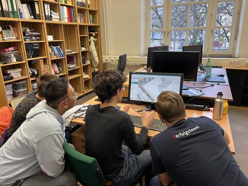 Vier Jugendliche arbeiten gemeinsam an einem Computer an einem Tisch in einer Bibliothek; der Bildschirm zeigt eine Architektur- bzw. Karten-Darstellung. Im Hintergrund stehen Bücherregale und Fenster.