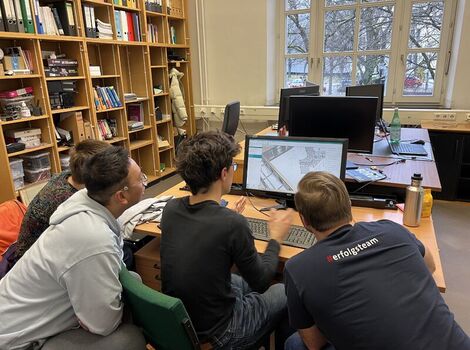 Vier Jugendliche arbeiten gemeinsam an einem Computer an einem Tisch in einer Bibliothek; der Bildschirm zeigt eine Architektur- bzw. Karten-Darstellung. Im Hintergrund stehen Bücherregale und Fenster.