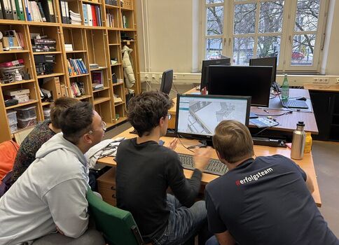 Vier Jugendliche arbeiten gemeinsam an einem Computer an einem Tisch in einer Bibliothek; der Bildschirm zeigt eine Architektur- bzw. Karten-Darstellung. Im Hintergrund stehen Bücherregale und Fenster.