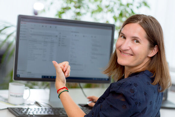 Eine Frau sitzt in einem Büro an einem Schreibtisch vor einem Computermonitor, lächelt in die Kamera und zeigt mit der Hand auf den Bildschirm.
