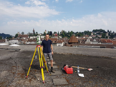 Un homme debout sur un toit, près d’un trépied de relevé et d’un instrument de mesure, avec une mallette et des outils posés au sol; ville aux toits rouges et clochers en arrière-plan.